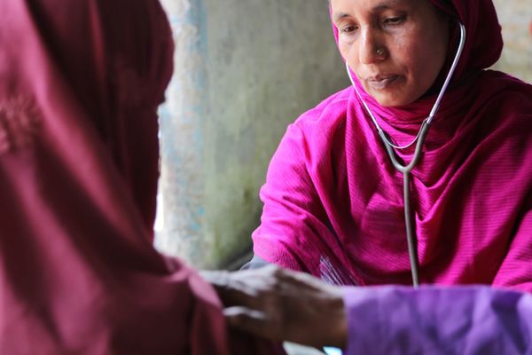 A woman in a pink top and head scarf visits with a female patient in Bangladesh. She wears a stethoscope.