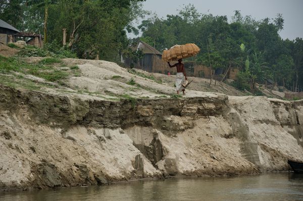 A man wearing a lungi balances bamboo fishing traps on his head while walking along the edge of a steep, eroded riverbank