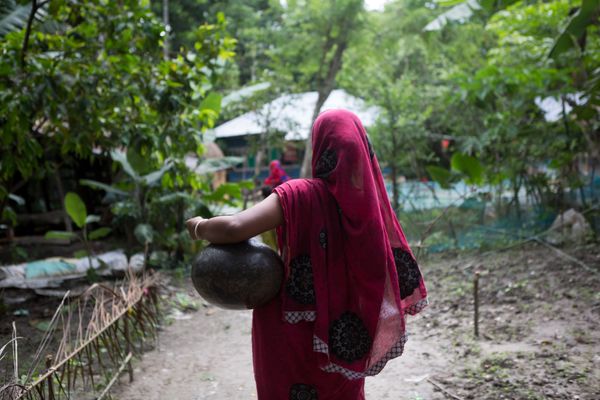 A woman wearing a pink sareer walks down a lane with a pot for water perched on her left hip