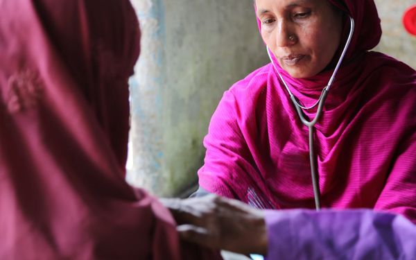 A woman in a pink top and head scarf visits with a female patient in Bangladesh. She wears a stethoscope.