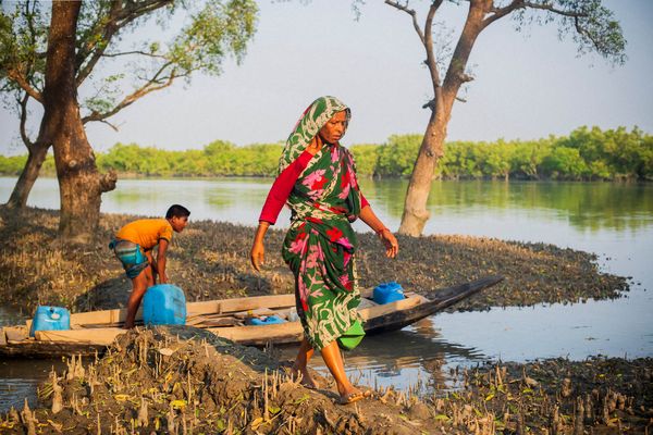 A woman wearing a green and pink saree walks carefully along a muddy river bank, away from a boy in a shallow boat who is preparing to collect water