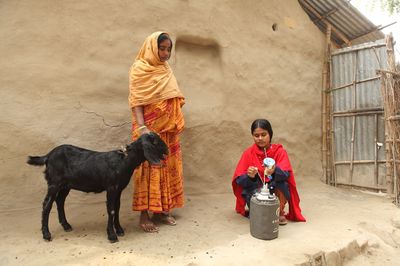 A woman with a red scarf crouches close to the container for her work and a woman with a goat looks on