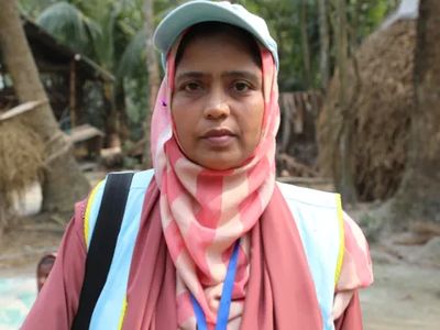 Shanaz Parbin Moni stands in a rural village. She wears a pale blue hat and a pink head scarf and top.