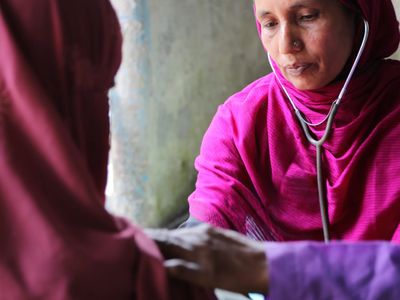 A woman in a pink top and head scarf visits with a female patient in Bangladesh. She wears a stethoscope.