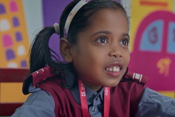 A young girl wearing a blue and maroon school uniform smiles as she sits in her colorful classroom