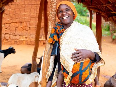 A woman wearting a blue and orange top with an orange head scarf smiles. There are black, white and brown goats in the background.