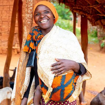 A woman wearting a blue and orange top with an orange head scarf smiles. There are black, white and brown goats in the background.