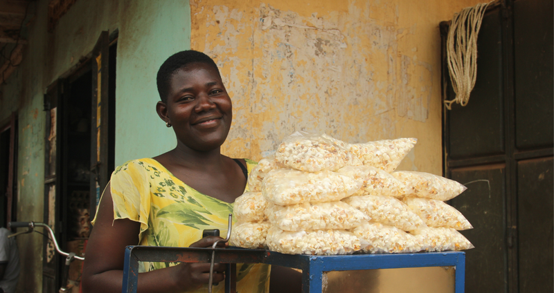 Juliet, a young woman in BRAC's youth empowerment program in Uganda, smiles as she poses behind her popcorn machine with a pile of bags of popcorn perched atop for sale.