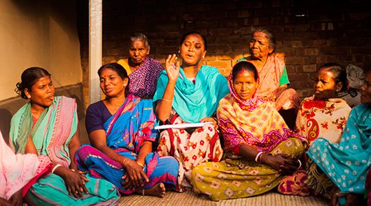 Eight women sit together. A woman with a blue scarf speaks to the group.