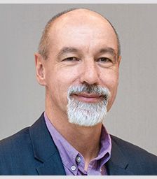 A man with a goatee smiles at the camera. He wears a purple dress shirt under a blue suit jacket.