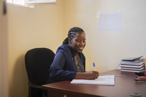 A woman wearing a blue suit jacket sits at a desk smiling as she works