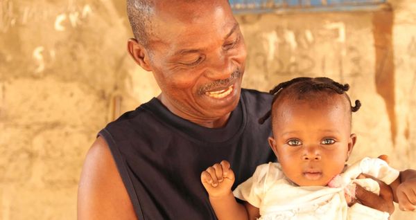 A Liberian father smiles as he looks down at his baby girl. He wears a black, sleeveless top. She wears a yellow dress.