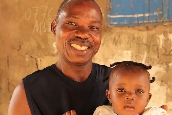 A Liberian father smiles as he holds his baby girl. He wears a black, sleeveless top. She wears a yellow dress.