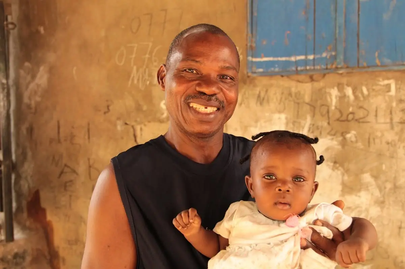 A Liberian grandfather smiles as he holds his baby granddaughter. He wears a black, sleeveless top. She wears a yellow dress.