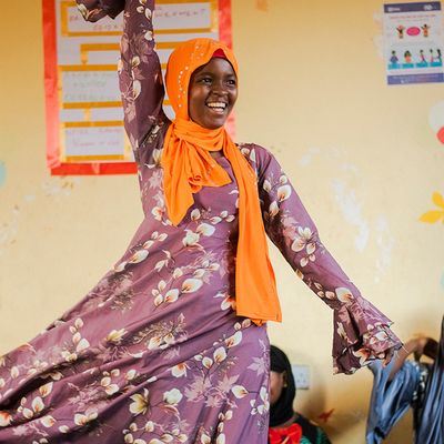A smiling girl dances while her peers watch, clap, and laugh in a girls' empowerment club in Tanzania.