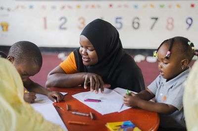 A woman is helping two young children draw at a small table in a classroom.