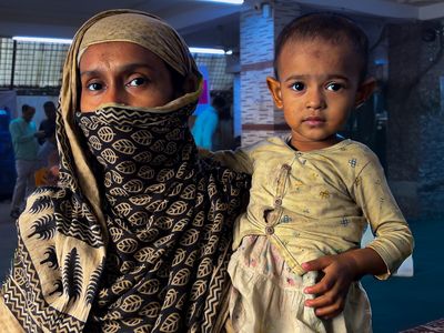 A woman wearing a blue scarf wtih beigh prints of petals looks at the camera while holding her baby who wears a yellow top