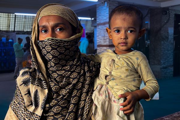 A woman wearing a blue scarf wtih beigh prints of petals looks at the camera while holding her baby who wears a yellow top