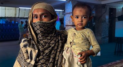 A woman wearing a blue scarf wtih beigh prints of petals looks at the camera while holding her baby who wears a yellow top