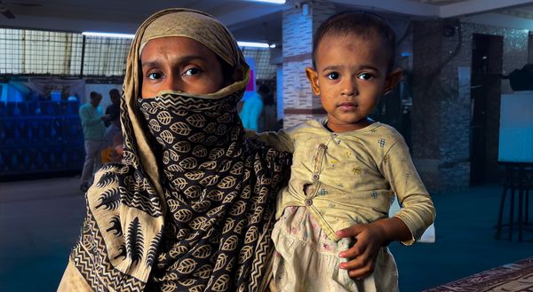 A woman wearing a blue scarf wtih beigh prints of petals looks at the camera while holding her baby who wears a yellow top