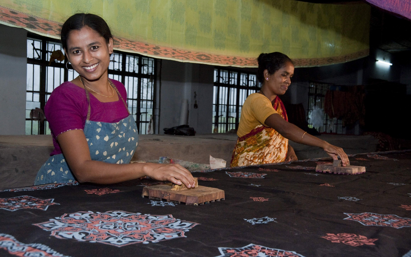 A woman poses and smiles while doing screen printing on a piece of fabric. She is an artisan behind Bangladesh's leading lifestyle brand, Aarong