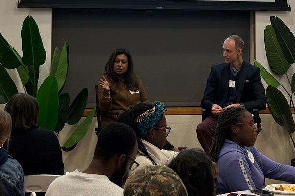 Tamara Hasan Abed wears a brown outfit and sits on the left of the stage. To her right is a man in a blue suit. Tamara gestures at the audience.