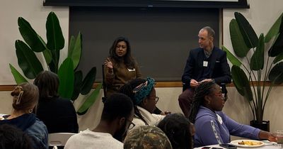 Tamara Hasan Abed wears a brown outfit and sits on the left of the stage. To her right is a man in a blue suit. Tamara gestures at the audience.