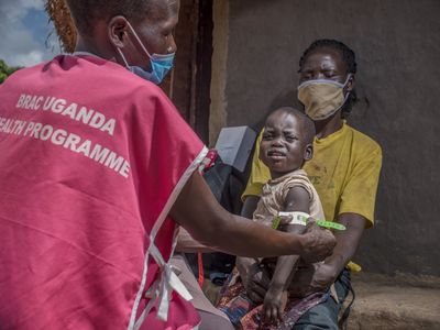 A health worker from the BRAC Uganda Health Program, wearing a mask, measures the mid-upper arm circumference of a young child to assess nutritional status. The child, sitting on a caregiver’s lap, looks distressed during the examination.