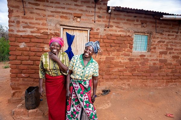 Two women in traditional attire stand in front of a brick house, showcasing cultural heritage and community.