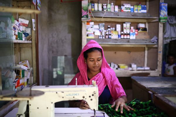 A woman wearing a pink head scarf sits at a sewing machine working on a green fabric