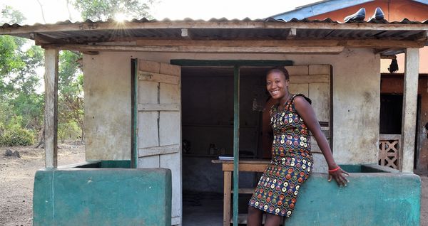 A woman stands in front of a small house, smiling, with a garden visible in the foreground.