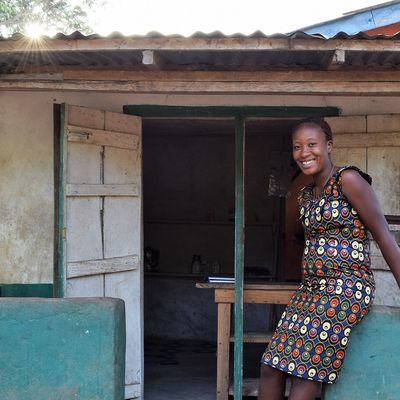 A woman stands in front of a small house, smiling, with a garden visible in the foreground.