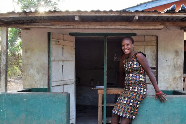 A woman stands in front of a small house, smiling, with a garden visible in the foreground.