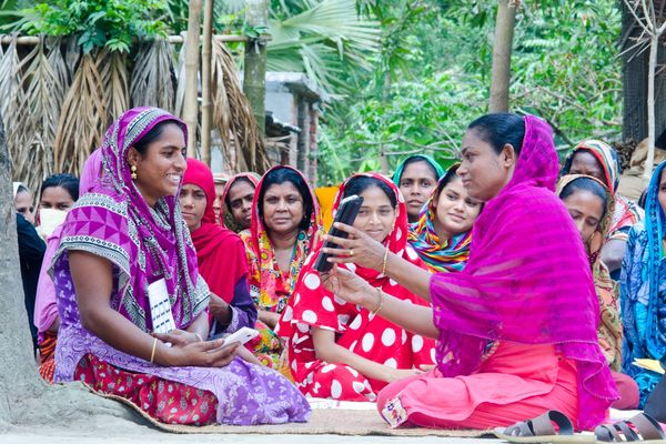 A group of women dressed in colorful sarees sit together outdoors, participating in a community meeting. Two women at the front are interacting using a smartphone.