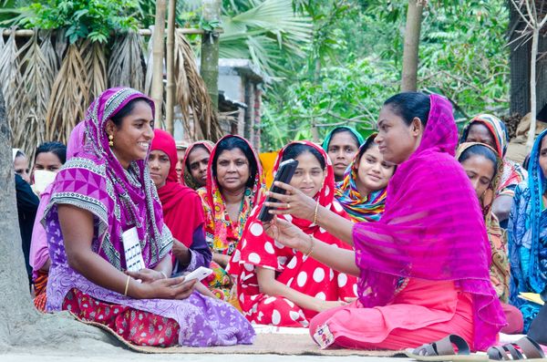 A group of women dressed in colorful sarees sit together outdoors, participating in a community meeting. Two women at the front are interacting using a smartphone.