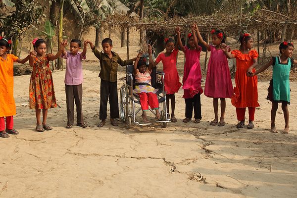 An image of children holding hands in a circle outside as part of a BRAC education program. One child included in the circle is in a wheelchair.