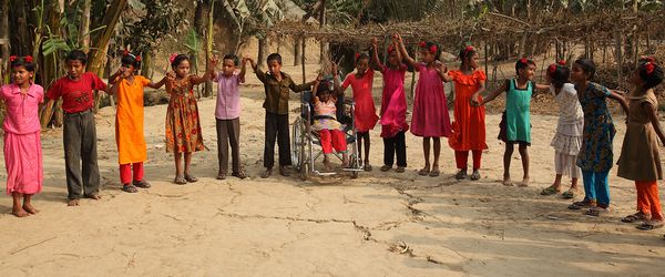 An image of children holding hands in a circle outside as part of a BRAC education program. One child included in the circle is in a wheelchair.
