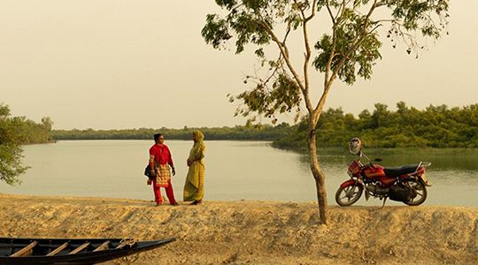 Two women stand in front of a lake talking.