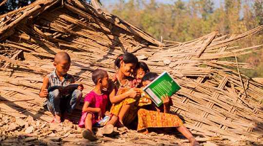 A woman sits on the remnants of a roof or wall, reading to a group of four children.