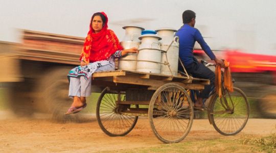 A man pedals a wooden van rickshaw in rural Bangladesh, carrying large metal milk containers while a woman in a red scarf and patterned dress sits on the side of the cart.