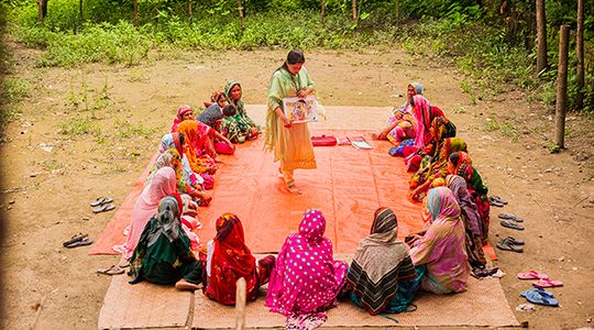 Over 15 women sit down in a circle and another woman stands in the middle.