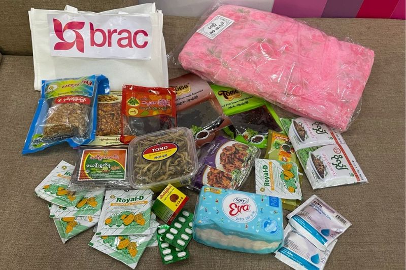 The items of an aid package are set out on a brown chair. The items include a pink mosquito net, sanitary pads, prepared food packets, and medicine.
