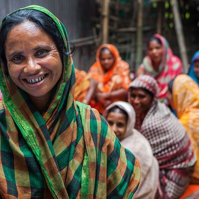 A group of Bangladeshi women, one stood smiling in the foreground, and at least 12 slightly blurred by depth of field in the background. All are wearing very colourful clothes of different patterns and hues. All are wearing headscarfs.